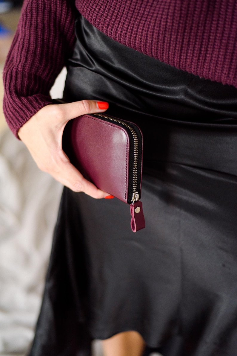 May include: A burgundy leather wallet with a zipper closure and a small wrist strap. The wallet is held against a black satin skirt. The person holding the wallet is wearing a burgundy sweater.