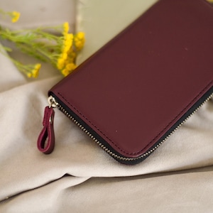 May include: A burgundy leather wallet with a black zipper and a matching pull tab. The wallet is rectangular and appears to be closed. The background includes a beige fabric and yellow flowers.