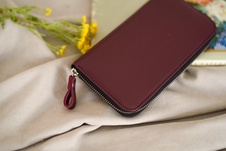 May include: A burgundy leather wallet with a black zipper and a matching pull tab. The wallet is rectangular and appears to be closed. The background includes a beige fabric and yellow flowers.