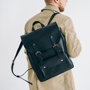 May include: A black leather backpack with a flap closure and adjustable straps. The backpack features gold-coloured buckles and a front pocket. The person in the image is wearing a beige trench coat.