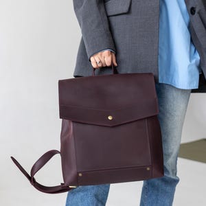 May include: A burgundy leather backpack with a flap closure and adjustable straps. The backpack has a rectangular shape with a top handle and gold-colored hardware. The person is wearing a gray blazer, a light blue shirt, and blue jeans.