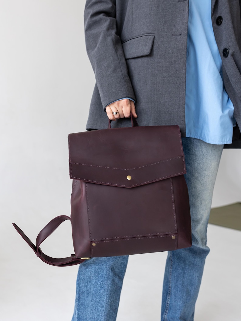 May include: A burgundy leather backpack with a flap closure and adjustable straps. The backpack has a rectangular shape with a top handle and gold-colored hardware. The person is wearing a gray blazer, a light blue shirt, and blue jeans.