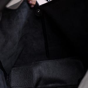 K&ouml;nnte beinhalten: Eine schwarze Ledertasche mit einer passenden Innentasche. Die Tasche ist offen und zeigt das Innere und ein hellrosa Smartphone, das hineingelegt wird. Das Leder hat eine strukturierte Oberfl&auml;che.