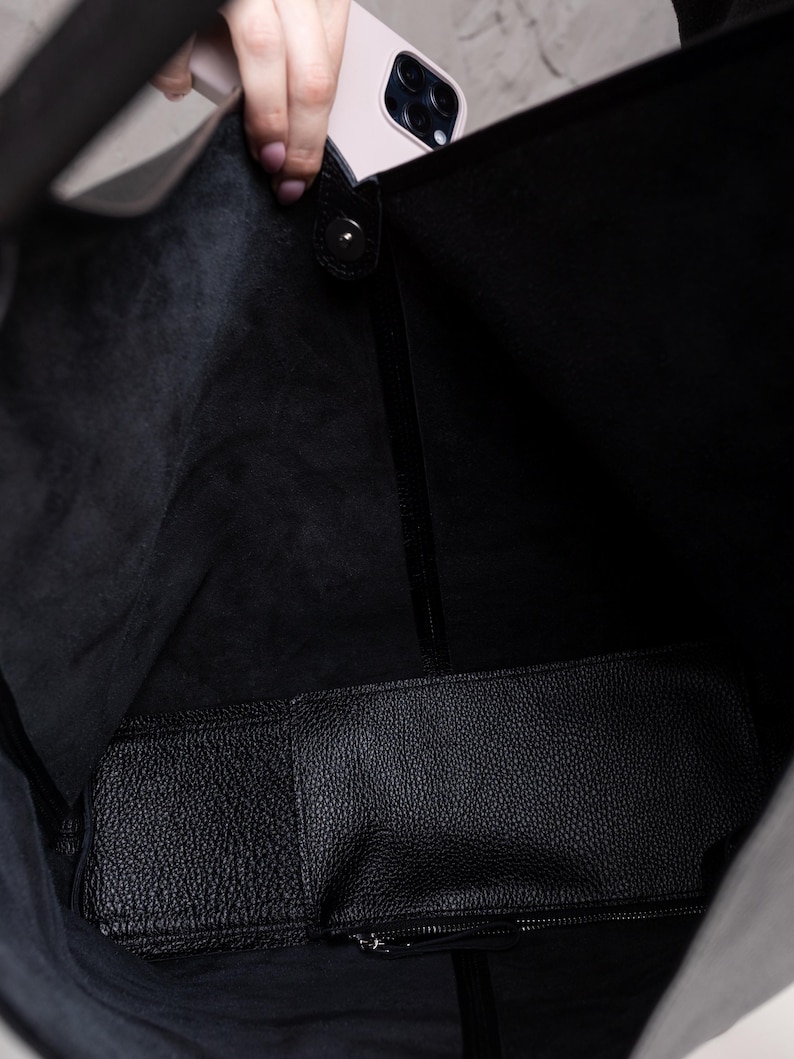 K&ouml;nnte beinhalten: Eine schwarze Ledertasche mit einer passenden Innentasche. Die Tasche ist offen und zeigt das Innere und ein hellrosa Smartphone, das hineingelegt wird. Das Leder hat eine strukturierte Oberfl&auml;che.