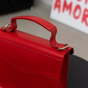 May include: A vibrant red handbag with a top handle and gold-colored hardware. The bag features a structured design with a flap closure and a small pocket on the front. The background includes a white canvas with the word "AMOR" in red.