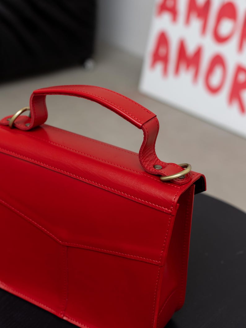 May include: A vibrant red handbag with a top handle and gold-colored hardware. The bag features a structured design with a flap closure and a small pocket on the front. The background includes a white canvas with the word "AMOR" in red.
