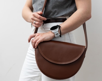 Puede incluir: Una mujer con una camiseta gris y pantalones vaqueros blancos lleva un bolso de hombro de cuero marrón en forma de media luna.