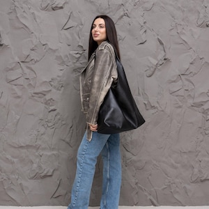 Puede incluir: Un bolso tote de cuero negro se lleva sobre el hombro. La persona lleva una chaqueta de cuero desgastada, vaqueros azul claro y bailarinas beige. El fondo es una pared gris texturizada.