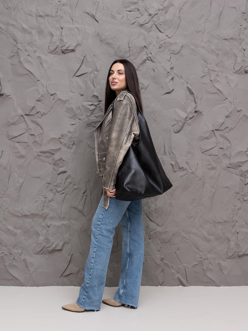 Puede incluir: Un bolso tote de cuero negro se lleva sobre el hombro. La persona lleva una chaqueta de cuero desgastada, vaqueros azul claro y bailarinas beige. El fondo es una pared gris texturizada.