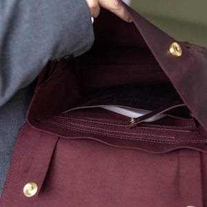 May include: Burgundy leather satchel bag with gold-colored hardware. The bag is open, revealing interior pockets and a flap closure. The bag is held open by a person.