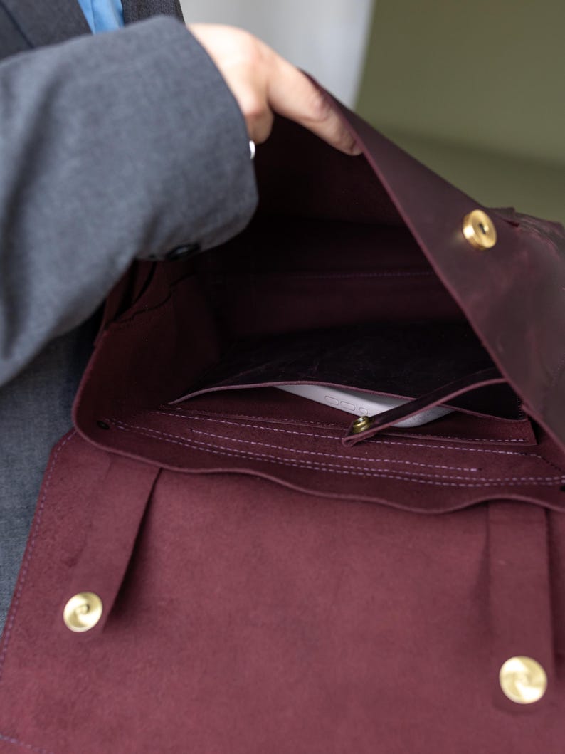 May include: Burgundy leather satchel bag with gold-colored hardware. The bag is open, revealing interior pockets and a flap closure. The bag is held open by a person.