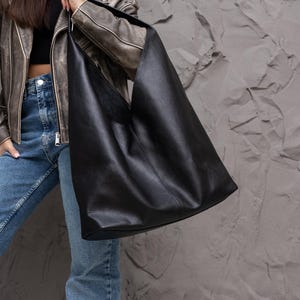 K&ouml;nnte beinhalten: Eine gro&szlig;e, schwarze Ledertasche mit weicher, strukturierter Optik. Die Tasche hat eine breite, offene Oberseite und einen einzelnen Schulterriemen. Die Tasche wird von einer Person gehalten, die eine braune Lederjacke und blaue Jeans tr&auml;gt.