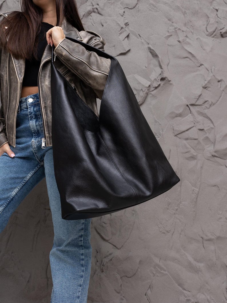 K&ouml;nnte beinhalten: Eine gro&szlig;e, schwarze Ledertasche mit weicher, strukturierter Optik. Die Tasche hat eine breite, offene Oberseite und einen einzelnen Schulterriemen. Die Tasche wird von einer Person gehalten, die eine braune Lederjacke und blaue Jeans tr&auml;gt.