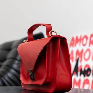 May include: A vibrant red leather handbag with a top handle and a flap closure. The bag features a rectangular shape with a structured design and a metal clasp. The bag is sitting on a black surface.