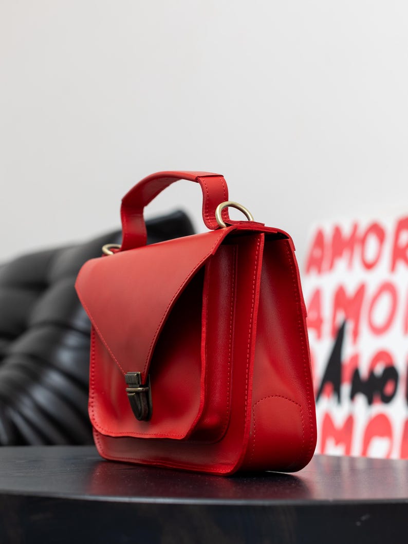 May include: A vibrant red leather handbag with a top handle and a flap closure. The bag features a rectangular shape with a structured design and a metal clasp. The bag is sitting on a black surface.