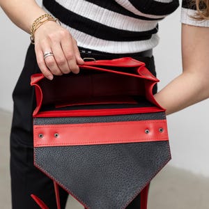 May include: A black and red leather handbag with a geometric design. The bag features a textured black front flap with a red accent stripe and interior. The bag has a silver clasp and red straps.