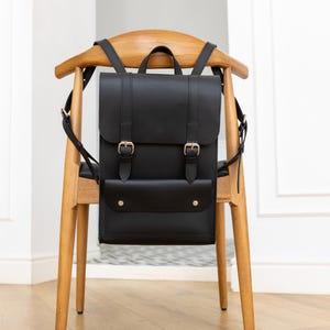 May include: A black leather backpack with a flap closure and gold-colored buckles. The backpack has a front pocket with two gold-colored rivets. The bag is hanging on a wooden chair with a curved back.