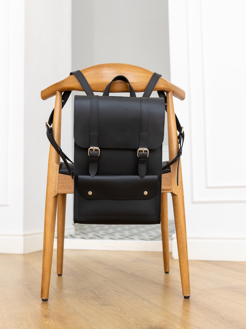 May include: A black leather backpack with a flap closure and gold-colored buckles. The backpack has a front pocket with two gold-colored rivets. The bag is hanging on a wooden chair with a curved back.