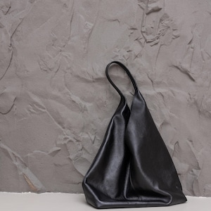 K&ouml;nnte beinhalten: Eine schwarze Ledertasche mit einem weichen, unstrukturierten Design. Die Tasche hat einen einzelnen Griff und eine glatte, matte Oberfl&auml;che. Die Tasche steht vor einem strukturierten grauen Hintergrund.