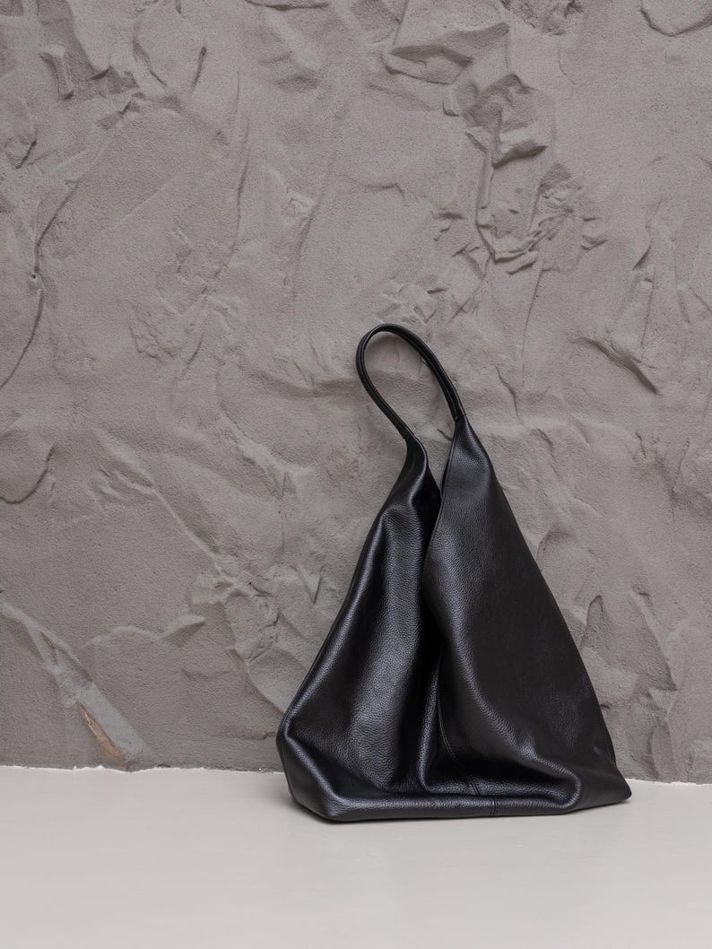 K&ouml;nnte beinhalten: Eine schwarze Ledertasche mit einem weichen, unstrukturierten Design. Die Tasche hat einen einzelnen Griff und eine glatte, matte Oberfl&auml;che. Die Tasche steht vor einem strukturierten grauen Hintergrund.