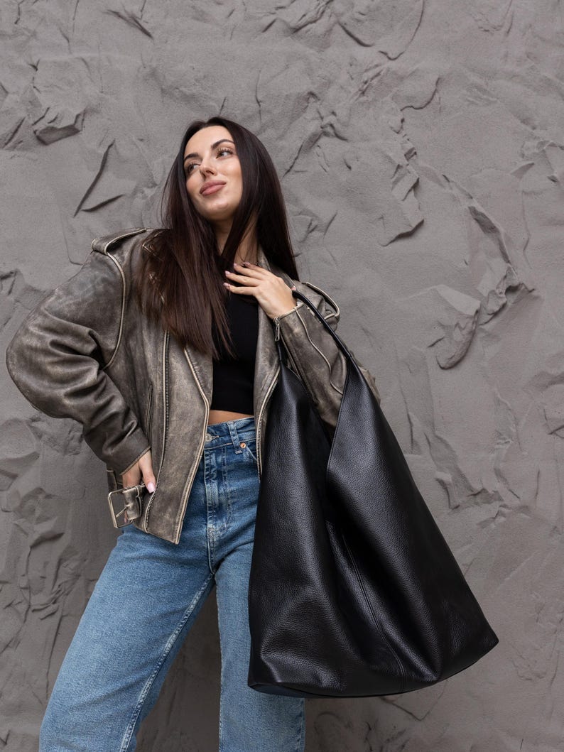 K&ouml;nnte beinhalten: Eine Frau tr&auml;gt eine gealterte braune Lederjacke, ein schwarzes Oberteil und blaue Jeans und h&auml;lt eine gro&szlig;e schwarze Leder-Tragetasche. Die Tasche hat ein schlichtes, unstrukturiertes Design. Der Hintergrund ist eine strukturierte graue Wand.