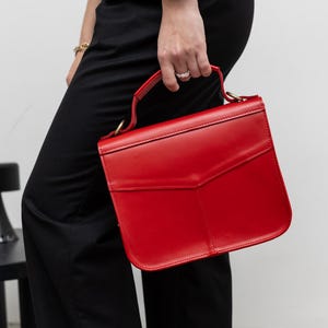 May include: A vibrant red handbag with a structured design. The bag features a top handle and a flap closure. The bag is held by a person wearing black pants and a watch.