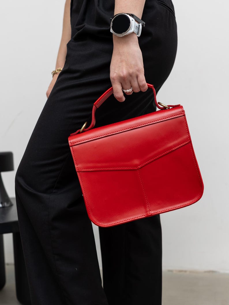 May include: A vibrant red handbag with a structured design. The bag features a top handle and a flap closure. The bag is held by a person wearing black pants and a watch.