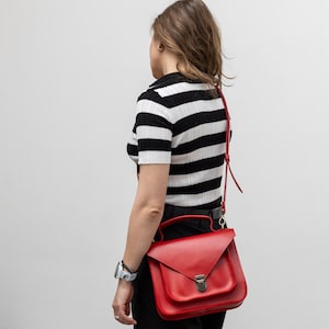 May include: A red leather satchel bag with a flap closure and a top handle. The bag has a shoulder strap and a metal clasp. The person is wearing a black and white striped shirt and black pants.