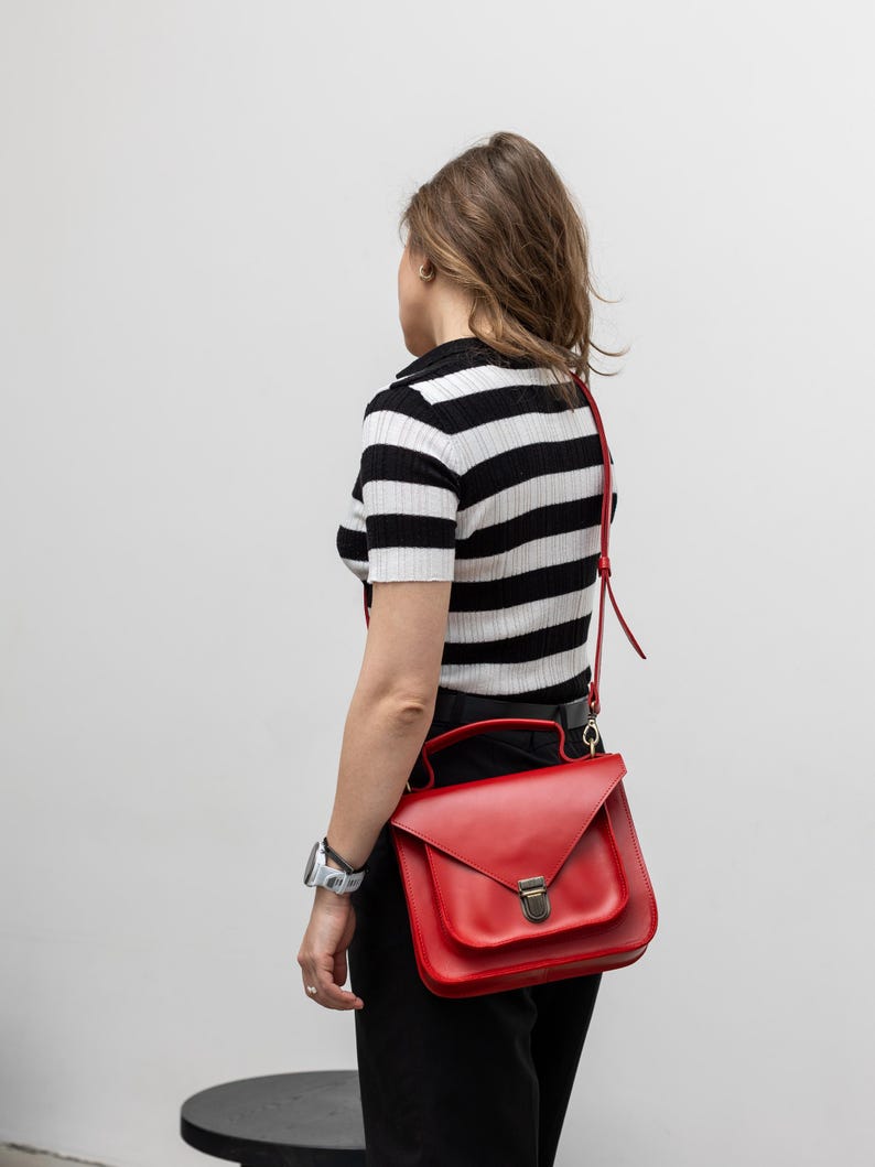 May include: A red leather satchel bag with a flap closure and a top handle. The bag has a shoulder strap and a metal clasp. The person is wearing a black and white striped shirt and black pants.