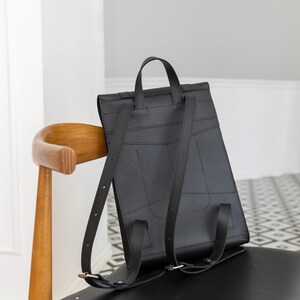 May include: A black leather backpack with a top handle and adjustable shoulder straps. The rectangular bag has a flap closure and is sitting on a chair. The bag is made of stitched panels and has a minimalist design.