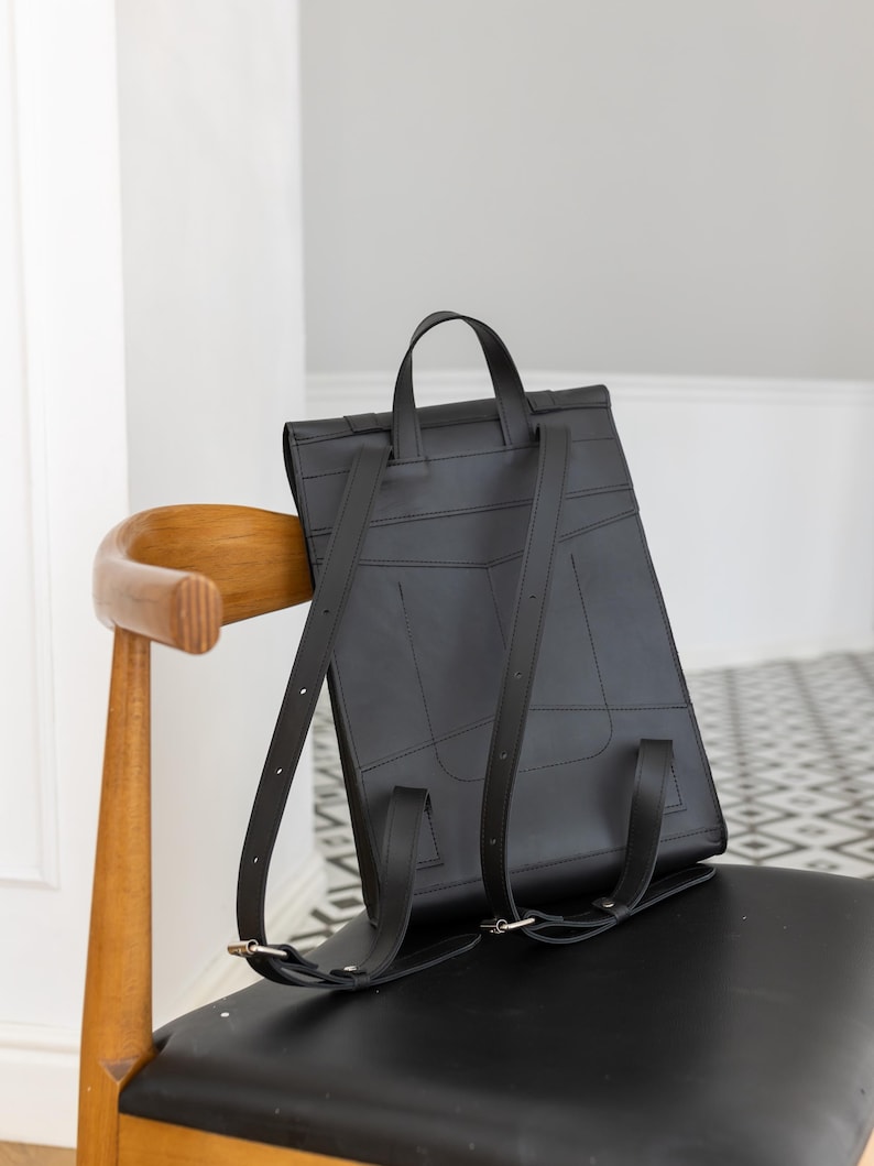 May include: A black leather backpack with a top handle and adjustable shoulder straps. The rectangular bag has a flap closure and is sitting on a chair. The bag is made of stitched panels and has a minimalist design.