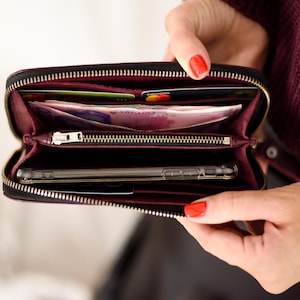 May include: Black zippered wallet with a burgundy interior, open to reveal cards, cash, and a smartphone. The wallet has a silver zipper and is held open, showcasing its contents. The overall theme is a practical and stylish accessory.
