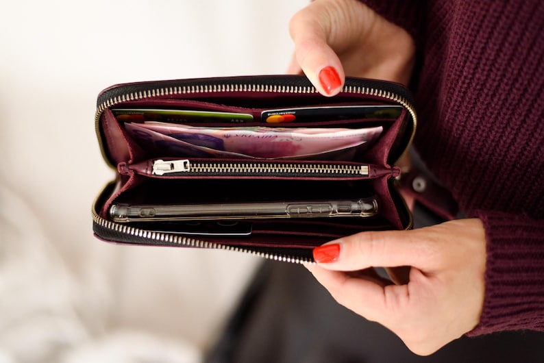May include: Black zippered wallet with a burgundy interior, open to reveal cards, cash, and a smartphone. The wallet has a silver zipper and is held open, showcasing its contents. The overall theme is a practical and stylish accessory.