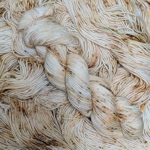 May include: Close-up of a skein of off-white yarn with brown and tan speckles. The yarn is tightly twisted and appears soft. The background is filled with more of the same yarn, creating a textured, tactile appearance. Ideal for knitting and crochet projects.