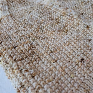 May include: Close-up of a hand-knitted item in cream and tan speckled yarn. The textured surface shows the detailed stitch work. A ball of yarn is visible in the background, suggesting a handmade craft project. The item appears to be a scarf or a similar garment.