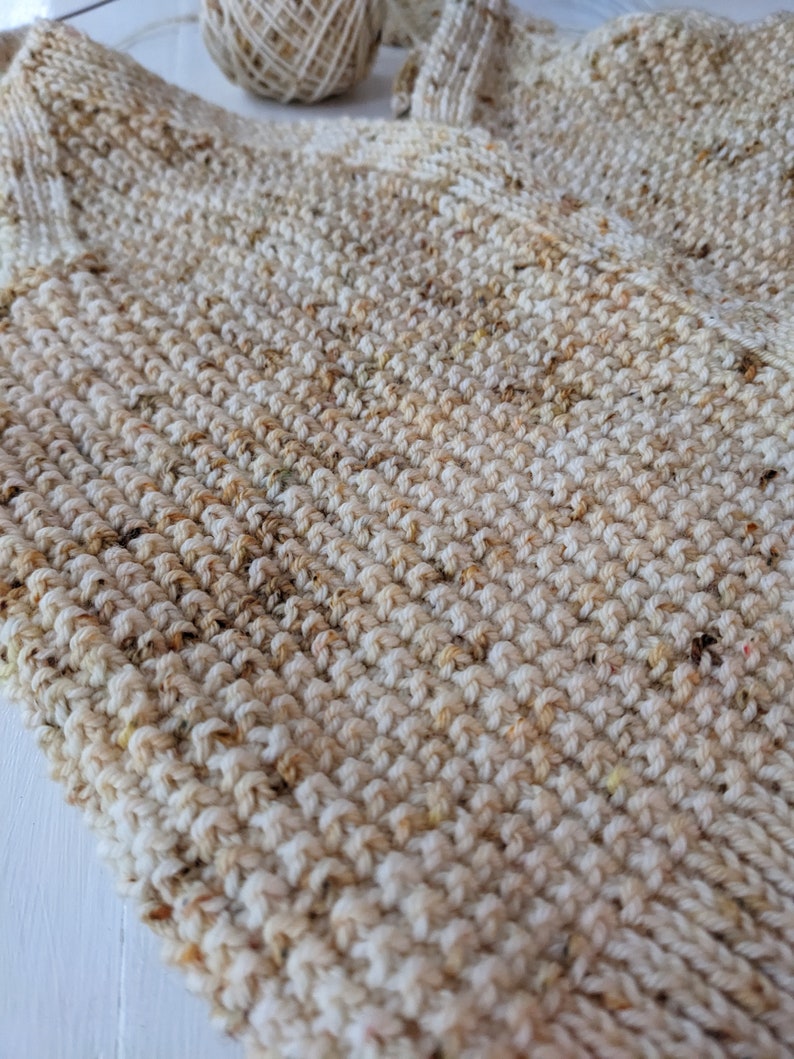 May include: Close-up of a hand-knitted item in cream and tan speckled yarn. The textured surface shows the detailed stitch work. A ball of yarn is visible in the background, suggesting a handmade craft project. The item appears to be a scarf or a similar garment.
