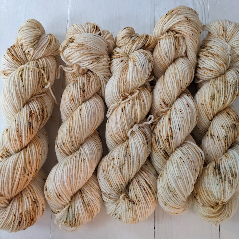 May include: Several skeins of off-white yarn with brown and green speckles. The yarn is tightly wound and appears soft, ideal for knitting or crocheting projects. The image is taken on a white wooden surface.