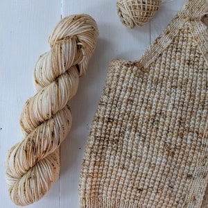 May include: A skein of cream-colored yarn with brown speckles, a small ball of yarn, and a knitted swatch. The yarn and knitted item are on a white surface. The yarn is likely for knitting or crocheting projects.
