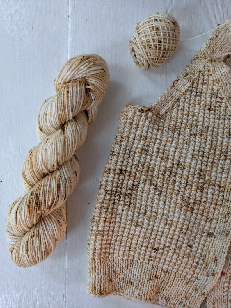 May include: A skein of cream-colored yarn with brown speckles, a small ball of yarn, and a knitted swatch. The yarn and knitted item are on a white surface. The yarn is likely for knitting or crocheting projects.