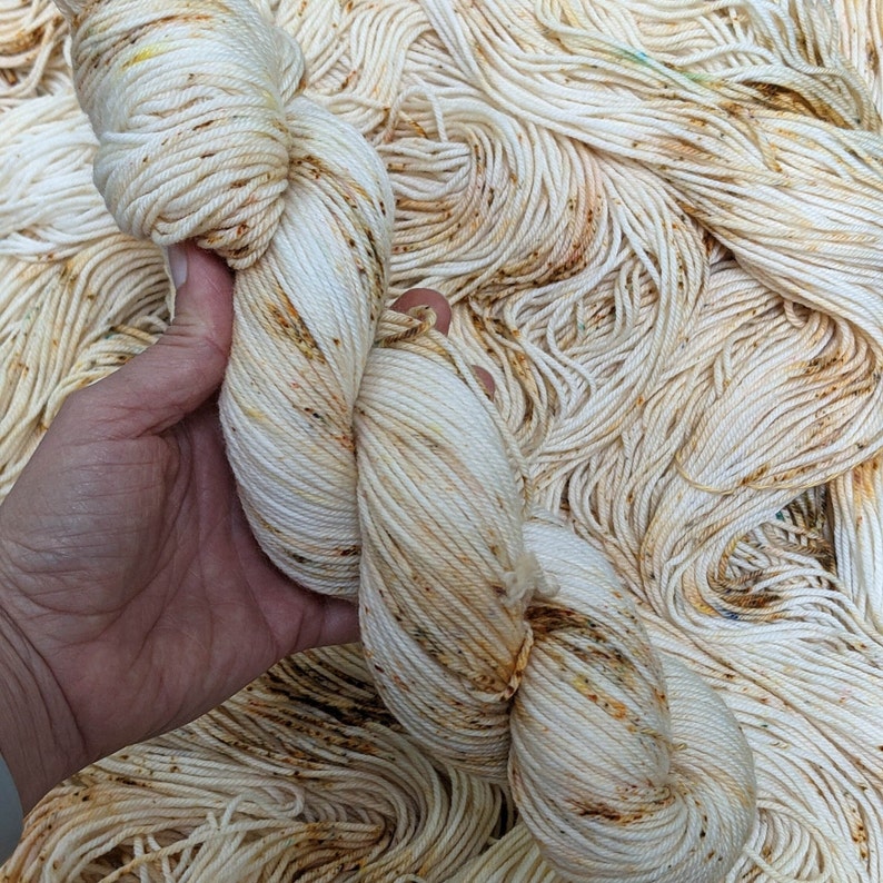 May include: A skein of off-white yarn with brown and yellow speckles is held in a hand. The yarn is part of a larger pile of similar yarn. The yarn appears to be soft and suitable for knitting or crocheting projects.