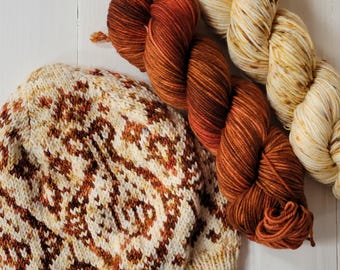 May include: A hand-knitted hat with a cream and brown pattern, next to two skeins of yarn. One skein is a gradient of orange and brown, and the other is cream with yellow and brown speckles. The items are on a white wooden surface.