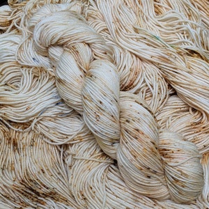 May include: Close-up of a skein of off-white yarn with brown and blue speckles. The yarn is tightly wound and appears soft, with a slightly textured surface. The background is filled with loose strands of the same yarn.