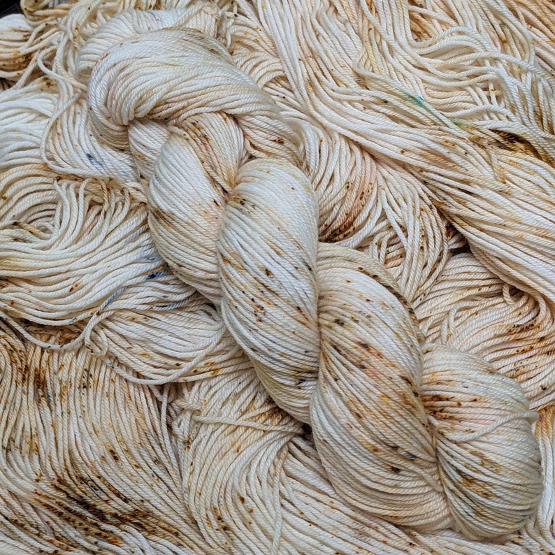 May include: Close-up of a skein of off-white yarn with brown and blue speckles. The yarn is tightly wound and appears soft, with a slightly textured surface. The background is filled with loose strands of the same yarn.