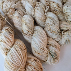 May include: Close-up of several skeins of off-white yarn with brown and tan speckles. The yarn is tightly wound and appears soft, ideal for knitting or crocheting projects. The texture of the yarn is clearly visible, highlighting its quality and potential for various crafts.