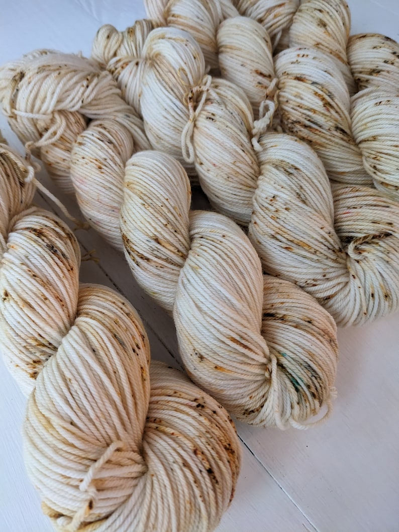 May include: Close-up of several skeins of off-white yarn with brown and tan speckles. The yarn is tightly wound and appears soft, ideal for knitting or crocheting projects. The texture of the yarn is clearly visible, highlighting its quality and potential for various crafts.