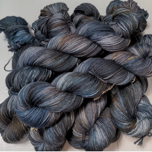 May include: A pile of hand-dyed yarn in shades of blue, gray, and brown. The yarn is twisted into skeins, showing variations in color and texture. The yarn appears soft and is suitable for knitting or crochet projects.
