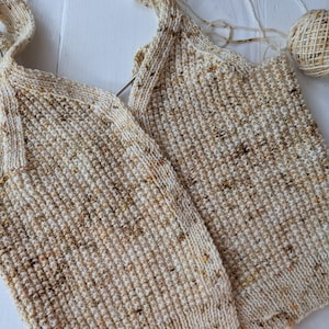 May include: Close-up of a partially completed knitted garment in cream and brown speckled yarn. The knitting needles are still in place, and a ball of yarn sits nearby. The textured stitch creates a cozy, handmade look.