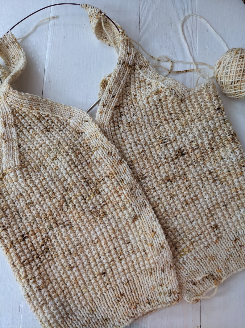 May include: Close-up of a partially completed knitted garment in cream and brown speckled yarn. The knitting needles are still in place, and a ball of yarn sits nearby. The textured stitch creates a cozy, handmade look.