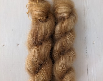 May include: Two skeins of light brown yarn, tightly wound into a twisted shape. The yarn has a soft, fuzzy texture and a slight sheen, with a few loose strands visible. The background is a white wooden surface.
