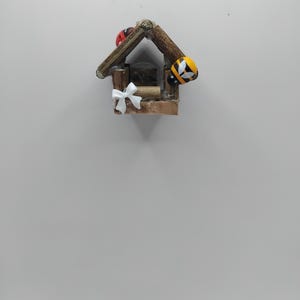 May include: A small wooden birdhouse with a brown roof and a white bow. The birdhouse is decorated with a red ladybird and a yellow and black bee. The birdhouse is a decorative item.